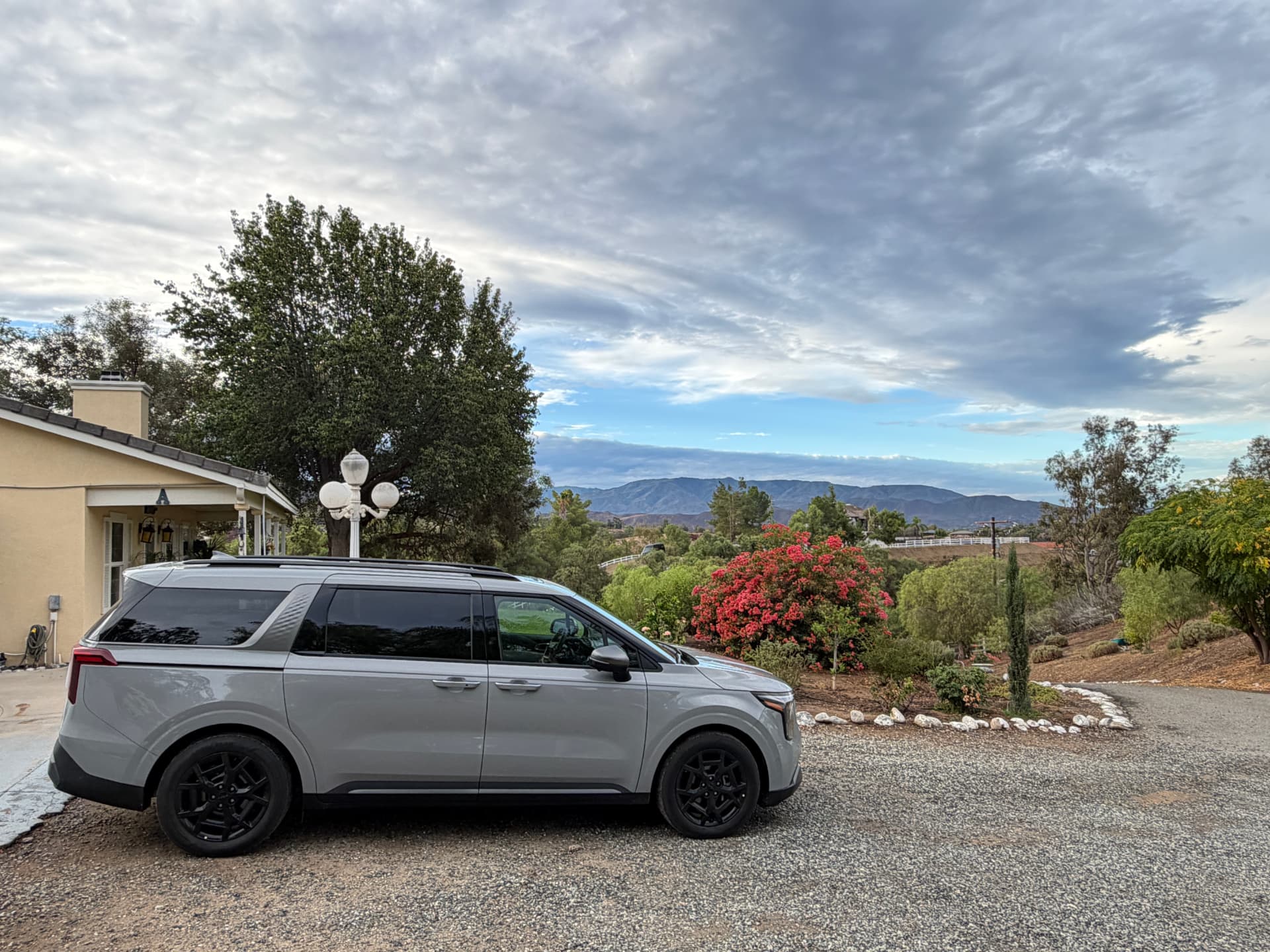 Silver Kia Carnival Hybrid parked beside a house with hills and a cloudy sky beyond